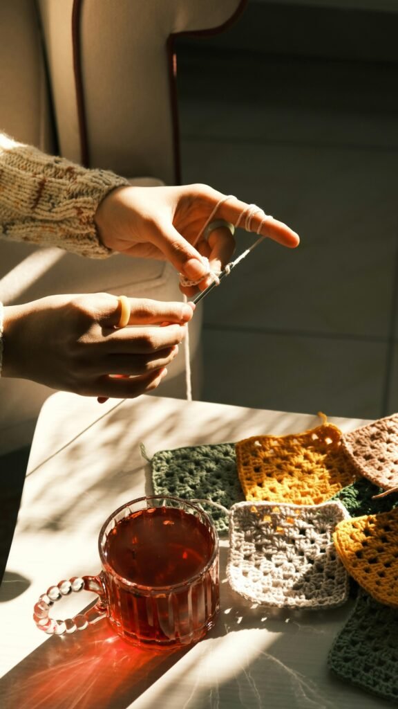 Two hands crocheting a granny square with colorful yarns, next to a cup of tea and soft wool balls on a cozy table.