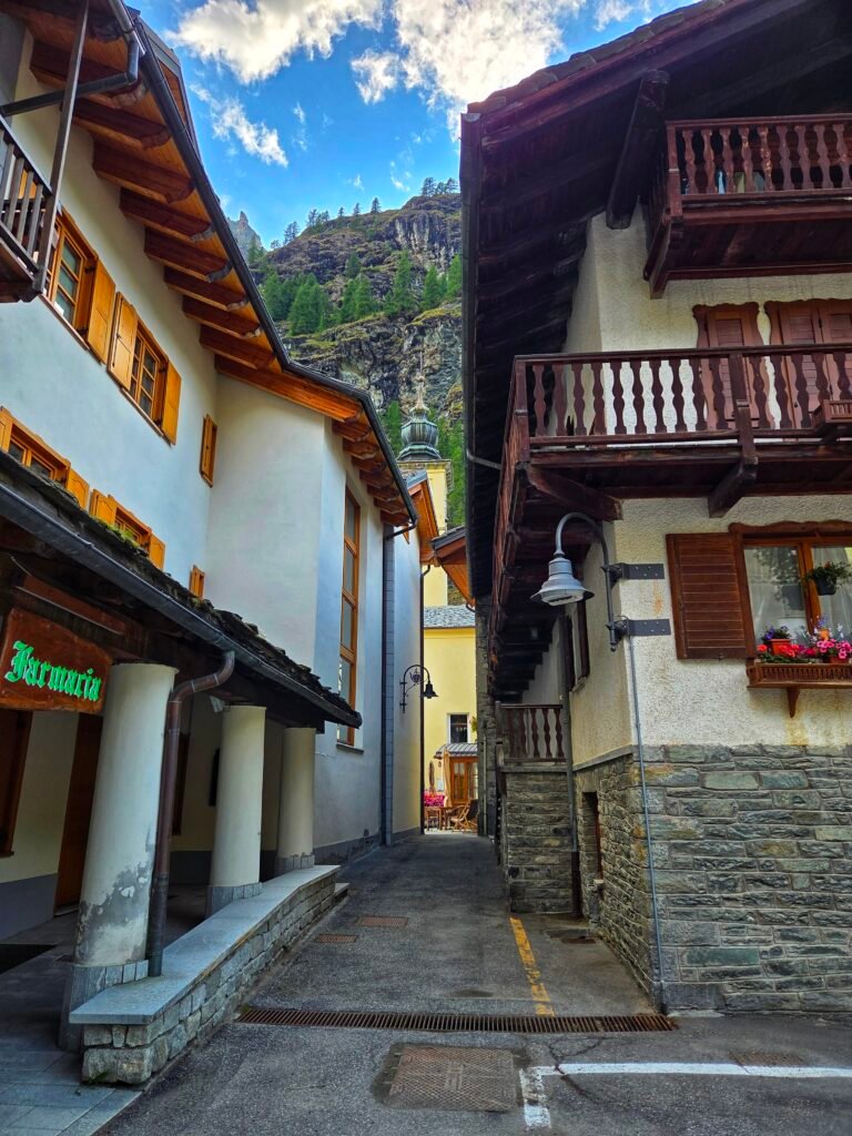Narrow alley between traditional alpine houses in a mountain village, with forested cliffs in the background.