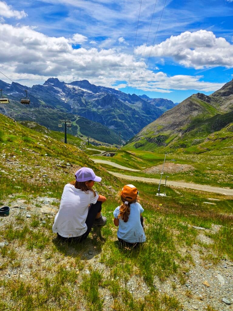 Two young girls sitting on a green meadow, looking out over a beautiful panoramic mountain valley.