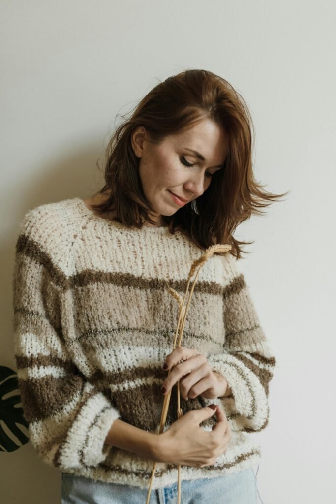 Woman wearing a handmade striped wool sweater in natural tones, gently holding dried wheat stems, standing against a neutral wall.