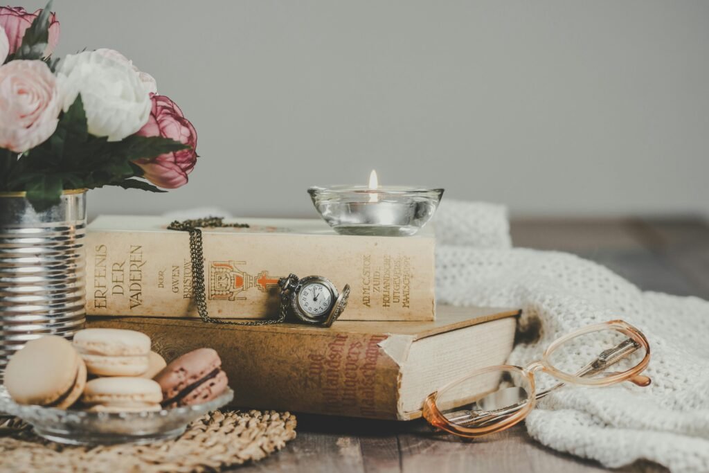 A wooden table with a vase of fresh flowers, books, a lit candle, an old-fashioned clock, eyeglasses, colorful macarons, and a soft blanket — all styled in a cozy, slow-living atmosphere.