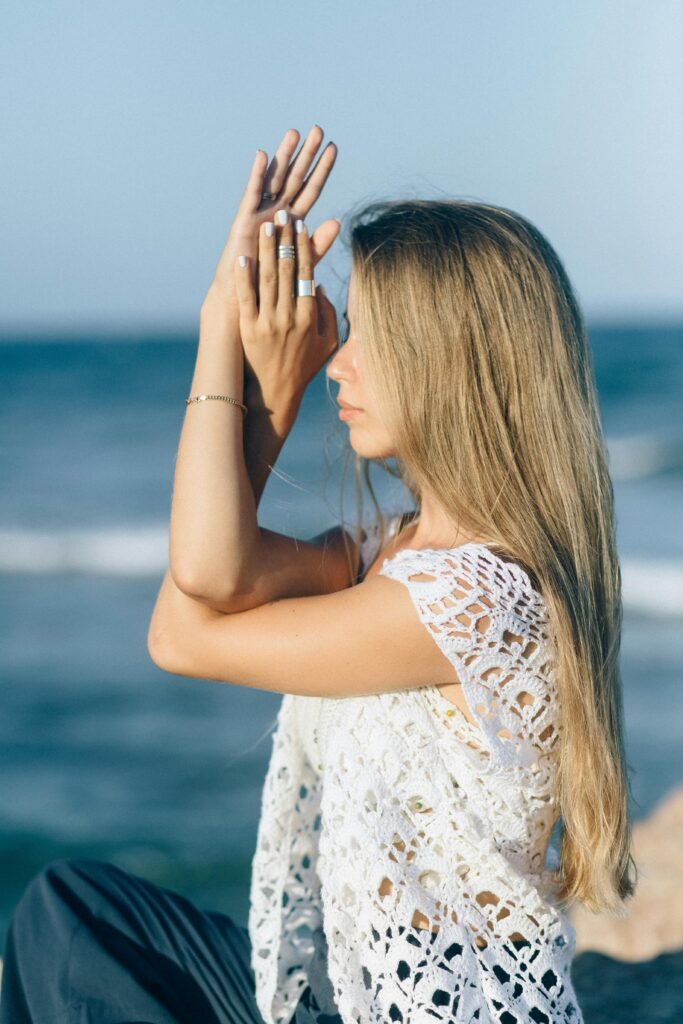 Young woman on the beach wearing a handmade crochet top, standing with arms crossed in a relaxed yoga pose.